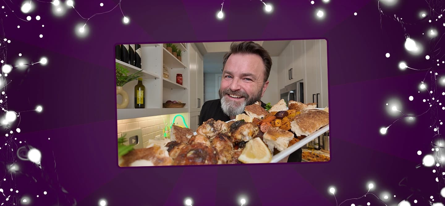 Smiling man holding a platter of roasted chicken and vegetables towards the camera in a modern kitchen.