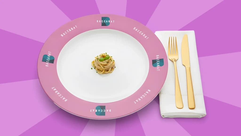 On a light purple background, a fork and a knife rest on a napkin to the right of a white plate with a purple border and a tiny portion of spaghetti. 