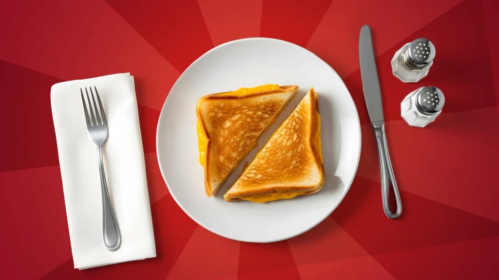 A fork rests on a white napkin, a plate with a grilled cheese is in the center, and a knife, salt, and pepper shaker are to the right. Everything is on a red background. 