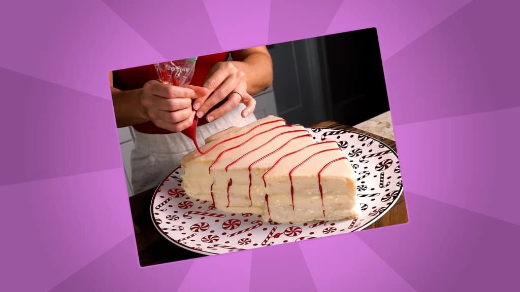 Close-up of a hand piping red icing in curved lines across the top of a Christmas tree-shaped cake on a holiday-patterned plate.
