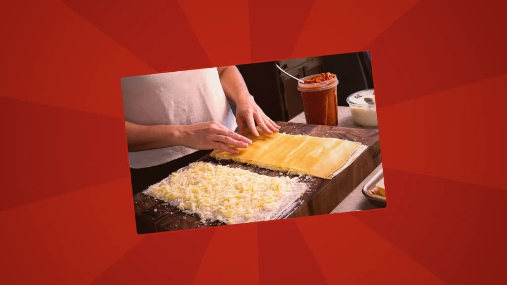 Hands spreading cheese evenly over layered lasagna sheets on a wooden cutting board.