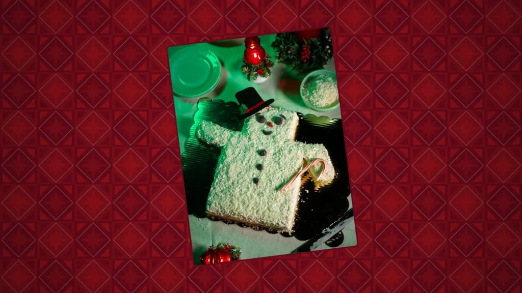A snowman-shaped coconut cake decorated with candy features, a paper top hat, and a candy cane, styled on a festive holiday table.
