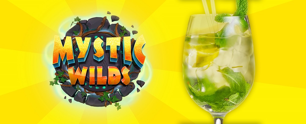 Against a two-toned yellow backdrop, the Cafe Casino 'Mystic Wilds' slots game logo is displayed on the left. The logo, in an orange and yellow cartoon font, is encircled by rocks and vines. To the right, a gin spritz is filled to the brim with ice and garnished with mint leaves, with two transparent plastic straws.