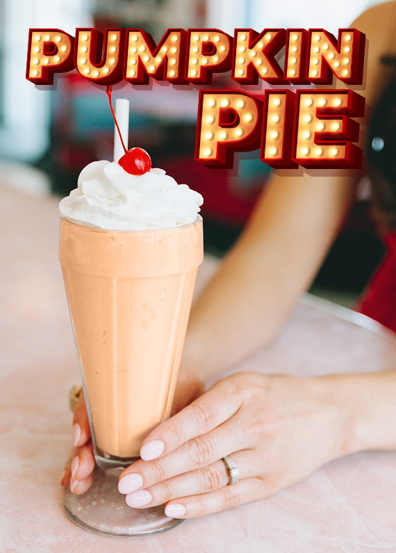 A tall pumpkin pie milkshake with cream and a cherry on top being placed onto a countertop by a young woman with the setting of a 50s diner in the background, featuring the words “Pumpkin Pie” in a Vegas-style sign