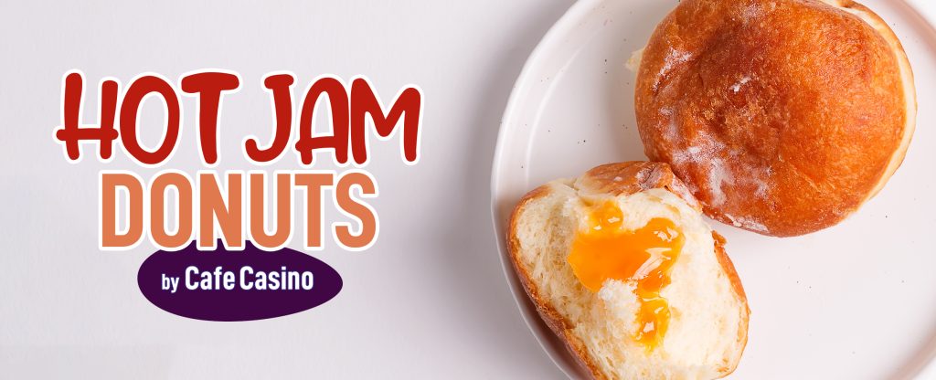 Two donuts sit on a white plate on a white table surface, one cut in half with cream and apricot jam. To the left, are words that read: “Hot Jam Donuts by Cafe Casino”.
