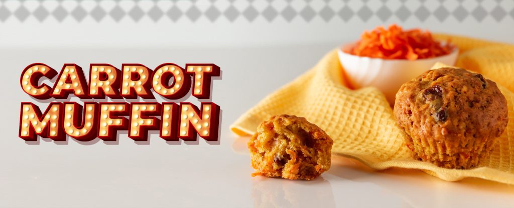 On top of a white high-gloss table surface sits a yellow tea towel, with a small white bowl filled with grated carrot, and in front, two carrot muffins – one large, one small. To the left is text in a Vegas-style font that reads “Carrot Muffin”.