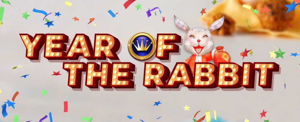 Prominently displayed in the middle of the image is text that reads “Year of the Rabbit” in Vegas-style font. Inside the O of “of” is a gold and blue crown symbol, representing Cafe Casino’s Hot Drop Jackpots. Behind the text is an animated rabbit character wearing a red jacket who’s holding up a red bag.