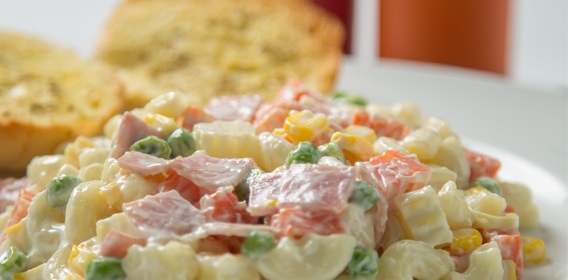 A white bowl filled with macaroni salad and a slightly blurred bread slice in the background.