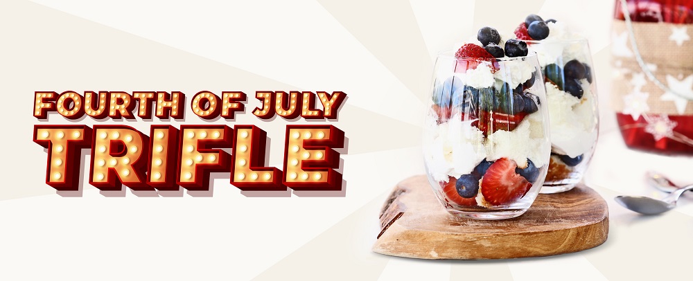We see two serving glasses on top of a wooden platter filled with trifles, in the fourth of July colors, featuring strawberries and blueberries surrounded by freshly whipped cream. To the right is a dessert spoon. To the left, we see a 3D-animated sign in the style of Vegas that reads ‘Fourth of July Trifle’, while behind, we see a striped white colored background.