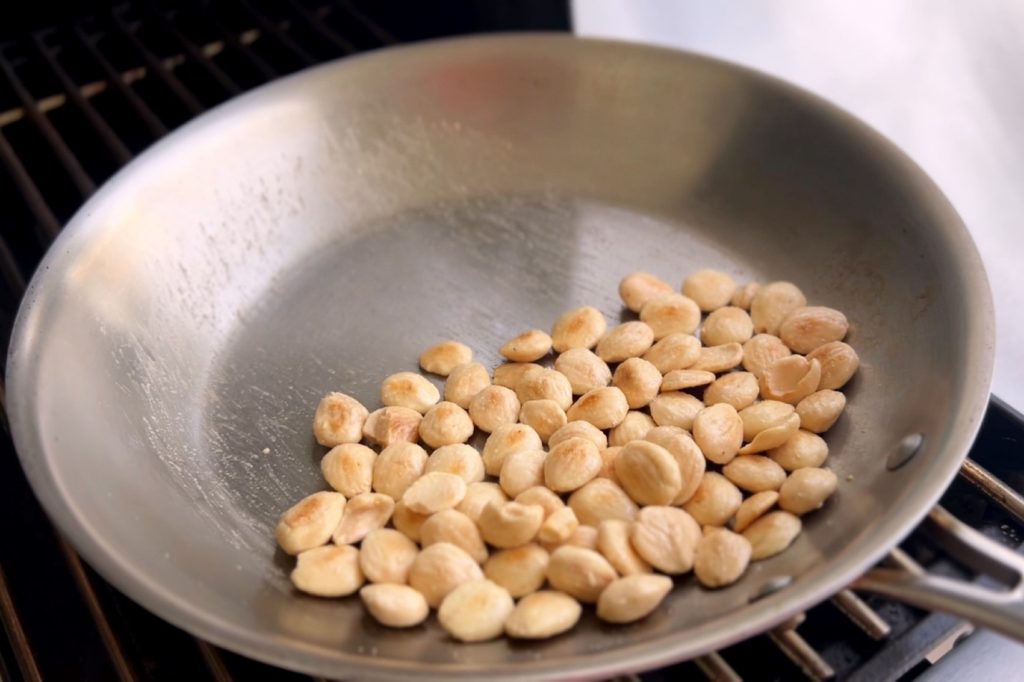 A large metal skillet sits on a barbecue grill, half full of almonds lightly roasting.