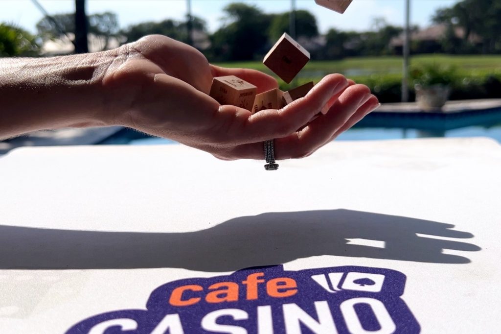 Cafe Casino influencer The Hunger Diaries hand is displayed holding dice, with a pool in the background and the Cafe logo in the foreground. 