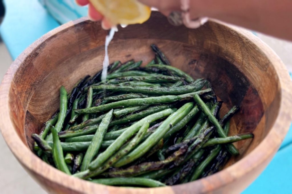 Green beans cooked in oil sit in a large wooden bowl, while the influencer The Hunger Diaries squeezes lemon juice from a fresh lemon overtop. The wooden bowl rests on a blue tablecloth on a table.