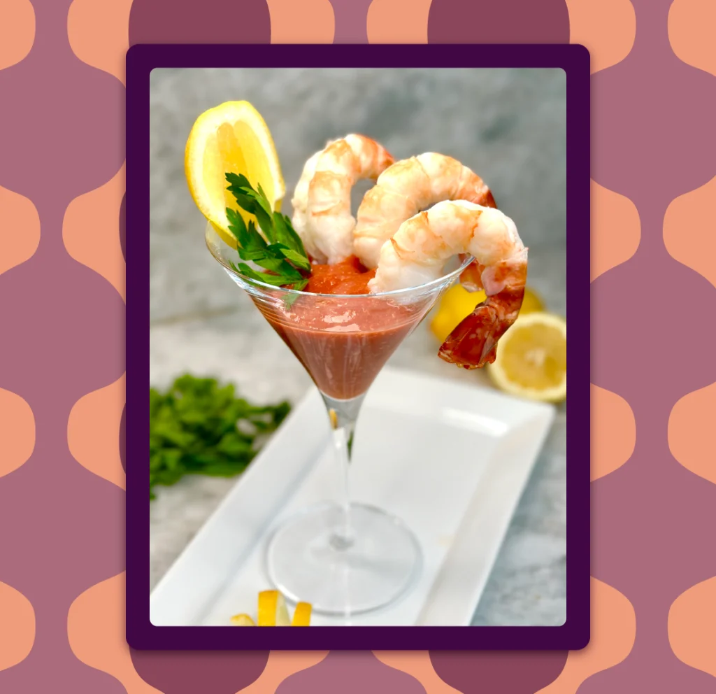 Three plump shrimp sit inside a classic cocktail glass, that’s filled with cocktail sauce; it sits on a white dish on a marble countertop.