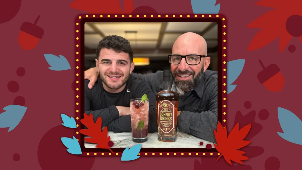 Father and Son known as Johnny Drinks stand together behind a cran-bourbon drink in a glass and a bottle of Johnny Drinks bourbon.