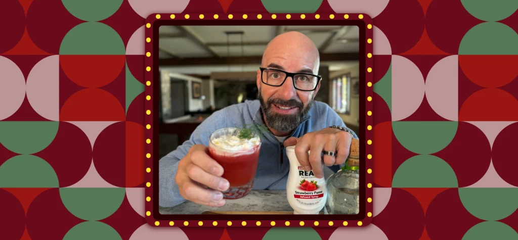 Johnny Drinks holds the Senor Santa cocktail in a tumbler glass, while holding a white bottle of strawberry pureé in his other hand; on a retro-style background.