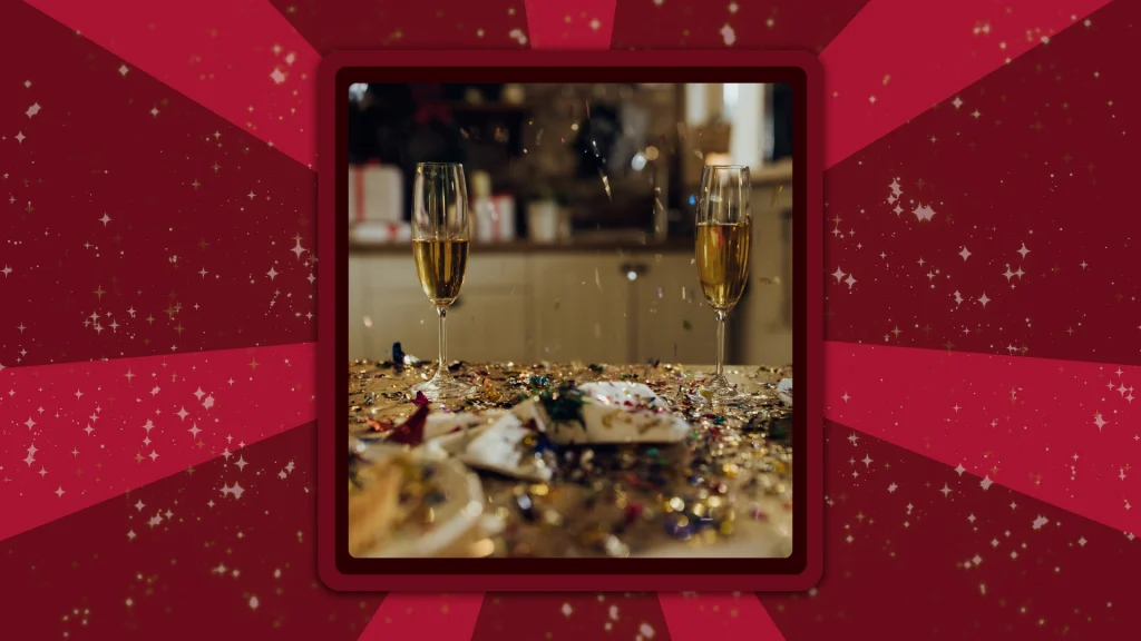 Two champagne flutes filled with champagne stand on a kitchen counter covered in New Year’s Eve sparkles and glitter.