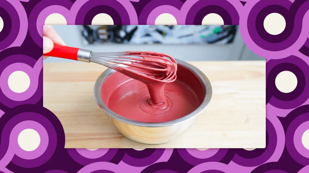 A bowl of pink red velvet lava cake batter, dripping off a whisk, on a wooden counter.