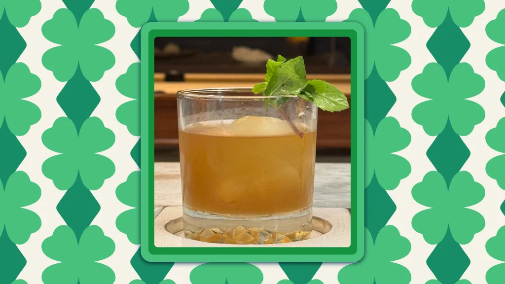 A Mint Old Fashioned in a glass with a piece of mint on the lip, on a green shamrock background.