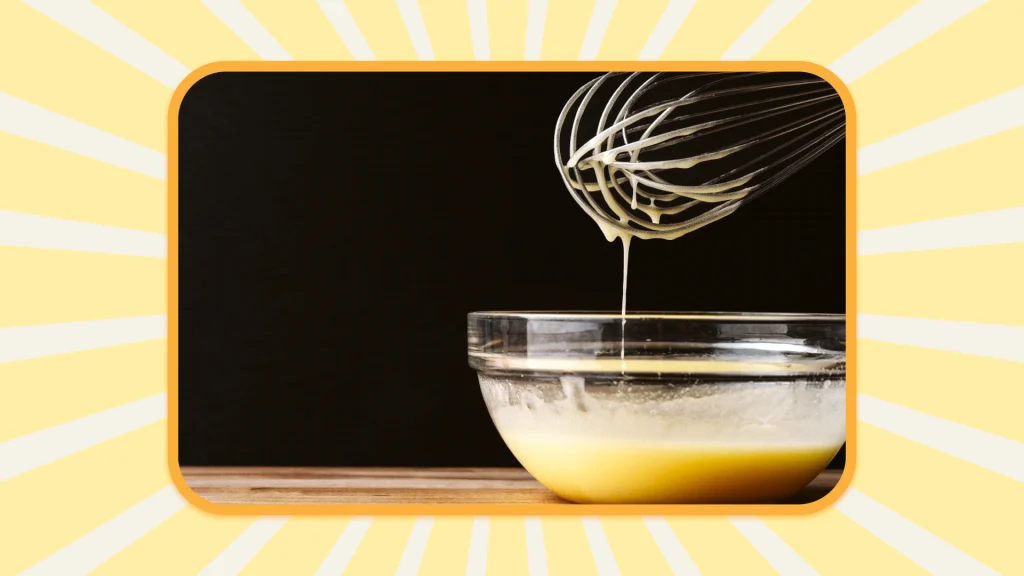 A bowl with pancake batter, with a whisk hovering above, dripping some of the batter.