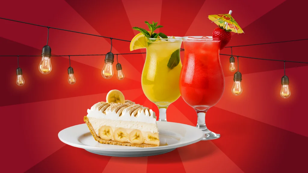 Two fruity cocktails stand to the right of a white plate with a slice of banana cream pie. Decorative lights dangle behind them on a red background.