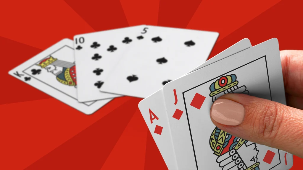 A hand holds an ace, and jack playing cards, and a king, 10, and 5 lay on a bright red background. 