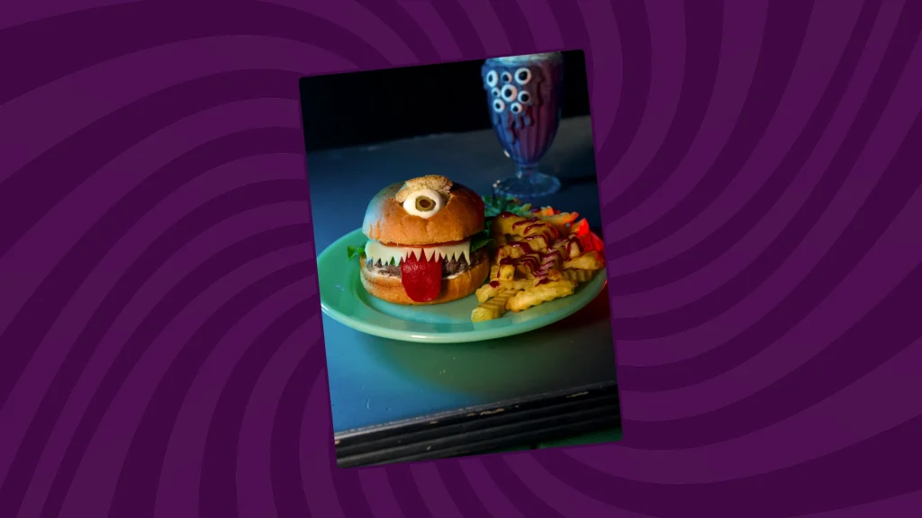 A burger that is made to look like a one-eyed monster is on a plate with fries and a shake in the background