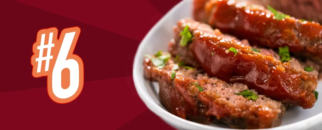 We see a white, glossy ceramic dish with three thick slices of meatloaf placed on top, sprinkled with freshly chopped parsley. To the left we see a “#6”, with an orange border. Behind is a dual-tone red and maroon striped background.